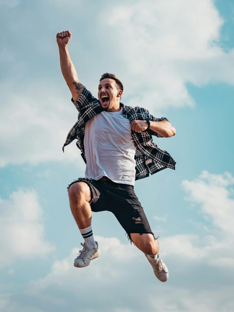 A joyful young man jumps midair with clouds and blue sky in the background, exuding energy and freedom.